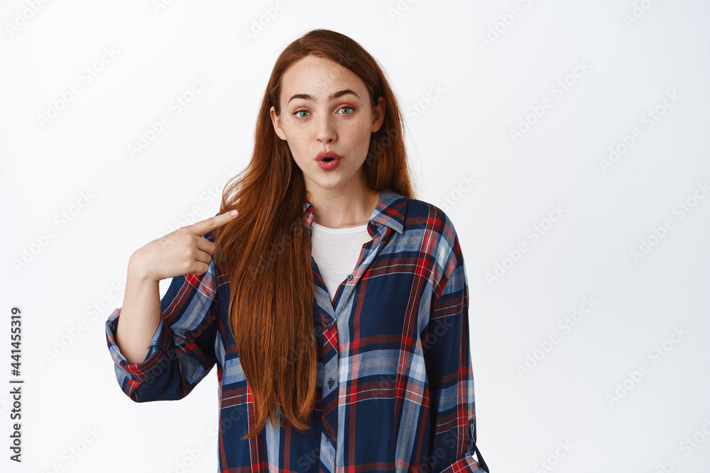 Image of redhead girl points at herself with surprised face, being chosen, winning something, look excited with disbelief, stands against white background Image of redhead girl points at herself with surprised face, being chosen, winning something, look excited with disbelief, stands against white background