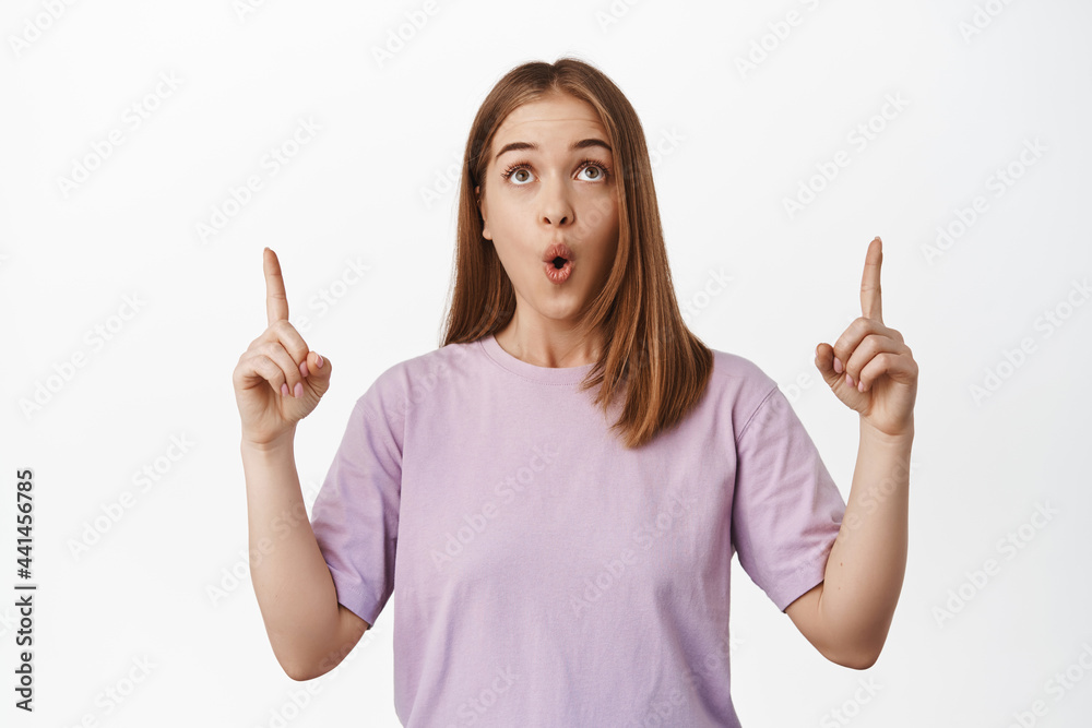 Image of surprised woman pointing and looking up with amazed face, say wow, checking out awesome advertisement, reading promotional text, standing over white background Image of surprised woman pointing and looking up with amazed face, say wow, checking out awesome advertisement, reading promotional text, standing over white background
