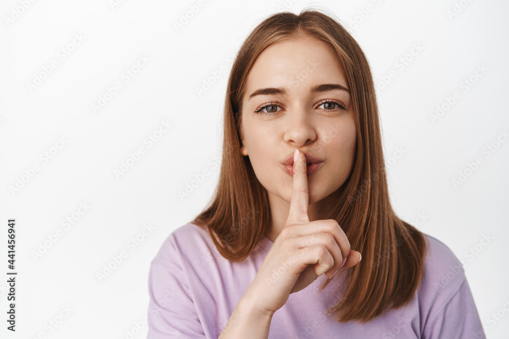 Hush. Close up portrait of blond girl shushing, smiling and asking to keep quiet, has secret, shhh gesture, press finger to lips, has surprise, standing against white background