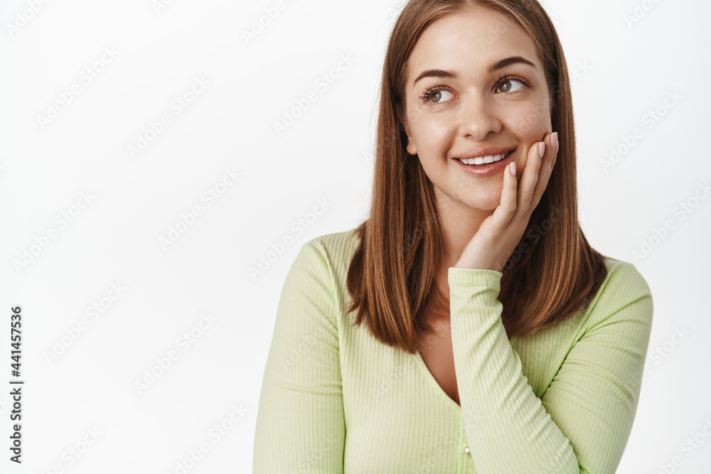 Close up of young woman smiling, touching clean glowing skin and gazing away at promotion text, daydreaming, concept of beauty and skincare, white background Close up of young woman smiling, touching clean glowing skin and gazing away at promotion text, daydreaming, concept of beauty and skincare, white background
