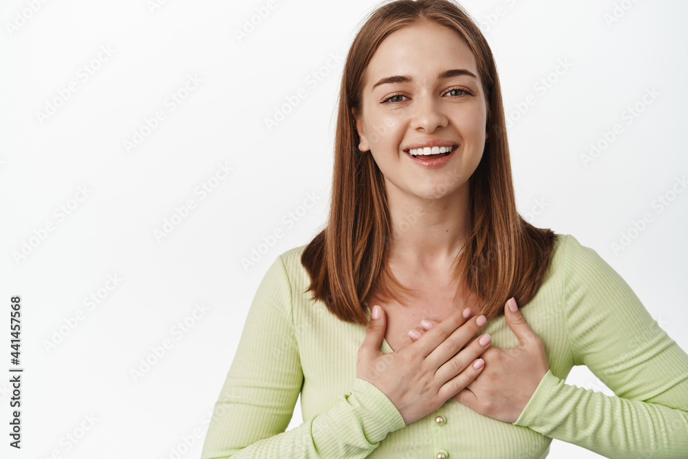 Portrait of touched girl say thank you, hold hands on heart and smiling pleased, receive praise, gift or compliment, standing against white background Portrait of touched girl say thank you, hold hands on heart and smiling pleased, receive praise, gift or compliment, standing against white background
