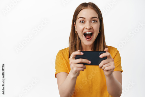Happy and thrilled young woman winning on mobile phone, holding smartphone both hands and scream excited, watching video stream on cellphone, white background