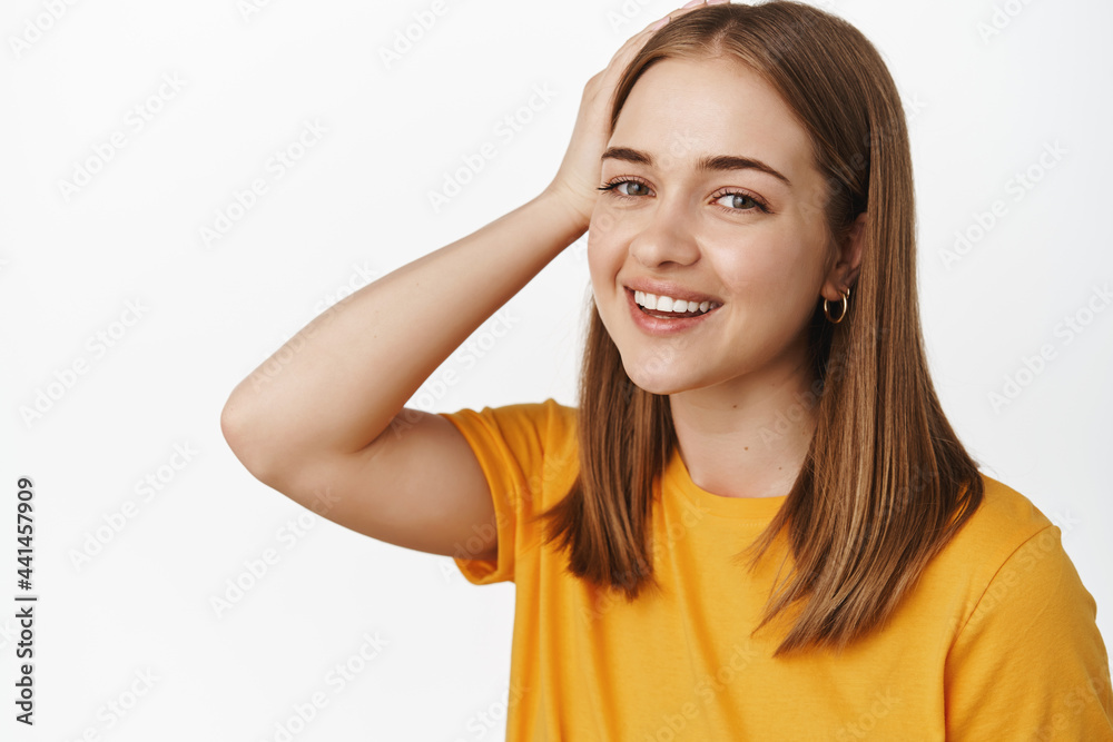 Close up portrait of beautiful candid girl, smiling happy, touching hairstyle and looking at camera carefree, standing in yellow t-shirt against white background Close up portrait of beautiful candid girl, smiling happy, touching hairstyle and looking at camera carefree, standing in yellow t-shirt against white background