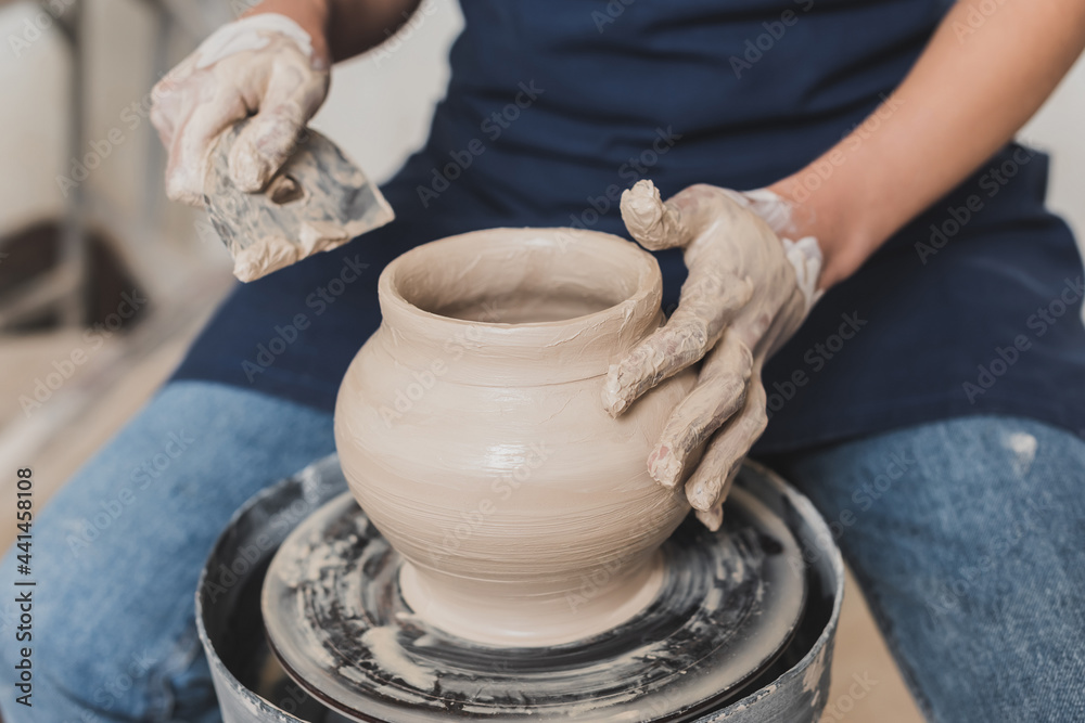 partial view of young african american woman in apron sitting on bench and shaping wet clay pot with scraper in pottery partial view of young african american woman in apron sitting on bench and shaping wet clay pot with scraper in pottery