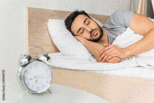 Young man sleeping near blurred alarm clock on bedside table