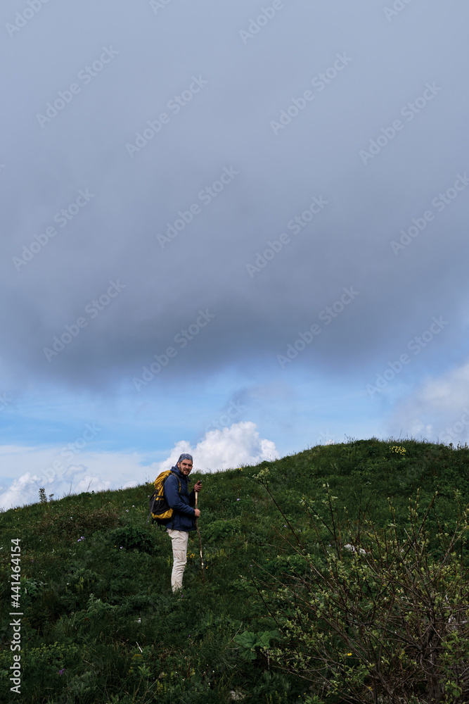 Male tourist with yellow large backpack travels together alone on plateau and studies new beauty of alpine meadows. Travel alone in mountains of Caucasus in summer. Male tourist with yellow large backpack travels together alone on plateau and studies new beauty of alpine meadows. Travel alone in mountains of Caucasus in summer.