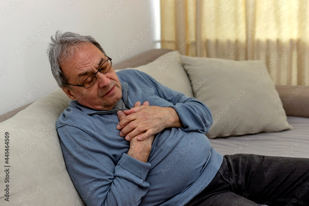 Worried senior man feeling unwell and having chest pain in the living room. Elderly man suffering from heart attack at home. Mature man feeling chest pain while sitting in the living room at home.