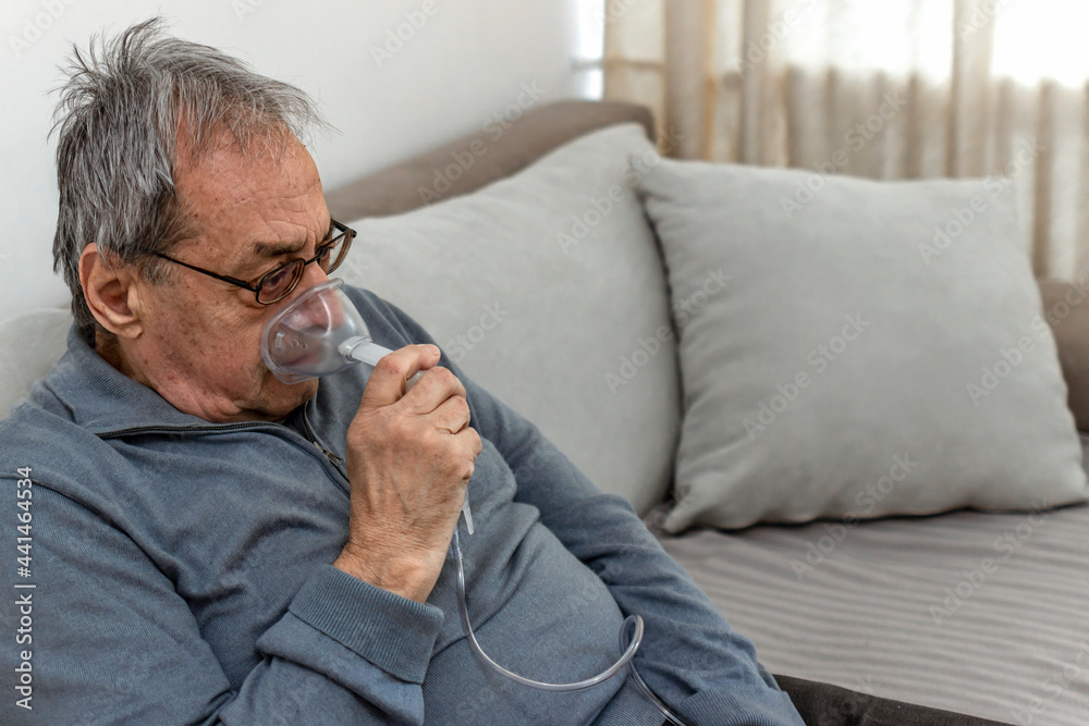 Portrait of elderly man with flu inhalation at home. The senior Caucasian man does inhalation with a nebulizer mask at home for coronavirus disease, sitting on couch at home alone. Health care concept Portrait of elderly man with flu inhalation at home. The senior Caucasian man does inhalation with a nebulizer mask at home for coronavirus disease, sitting on couch at home alone. Health care concept