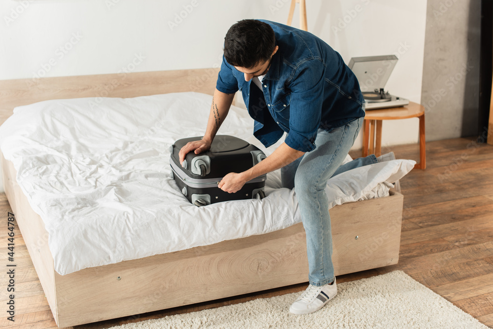 Young man zipping suitcase on bed at home