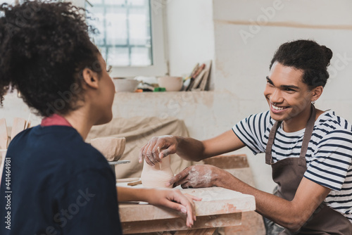 smiling young african american man sculpting clay pot and stick and looking at woman nearby in pottery