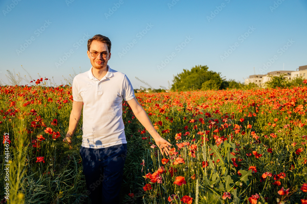 Portrait of young man in white polo standing in blooming red poppy field at sunset, Smiling boy in shirt and glasses confident and feeling freedom among flowers, sunny summer day, scarlet vivid poppy