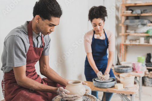 concentrated young african american couple in casual clothes and aprons shaping wet clay pots in pottery