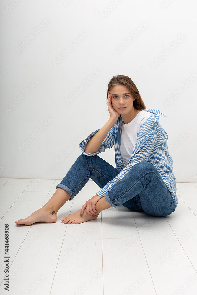 portrait of young attractive caucasian woman with long brown hair in t-shirt and blue jeans isolated on white background. skinny pretty female sitting on floor at studio. model tests of beautiful lady