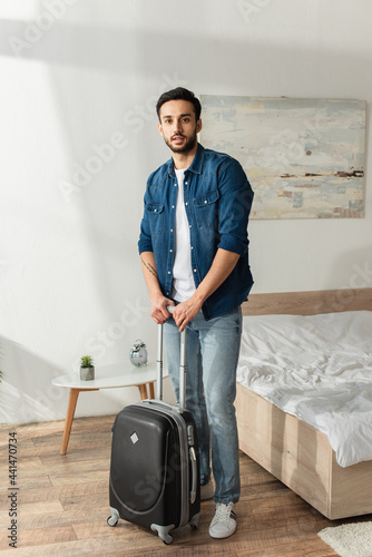 Young man with suitcase looking at camera in bedroom