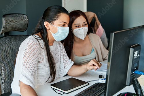 two young latina business women talking and working together in the office, wearing covid-19 protective masks in times of pandemic, teamwork concept