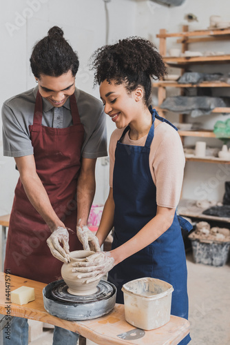 positive young african american couple in aprons shaping wet clay pot on wheel with hands in pottery