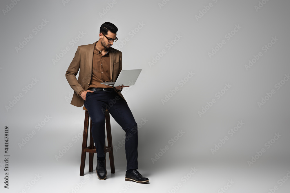 Arabian businessman looking at laptop and sitting on chair on grey background Arabian businessman looking at laptop and sitting on chair on grey background