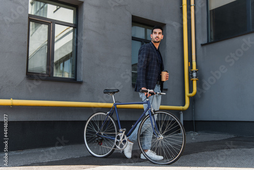 Young arabian businessman in jacket walking near bike and holding coffee to go outdoors
