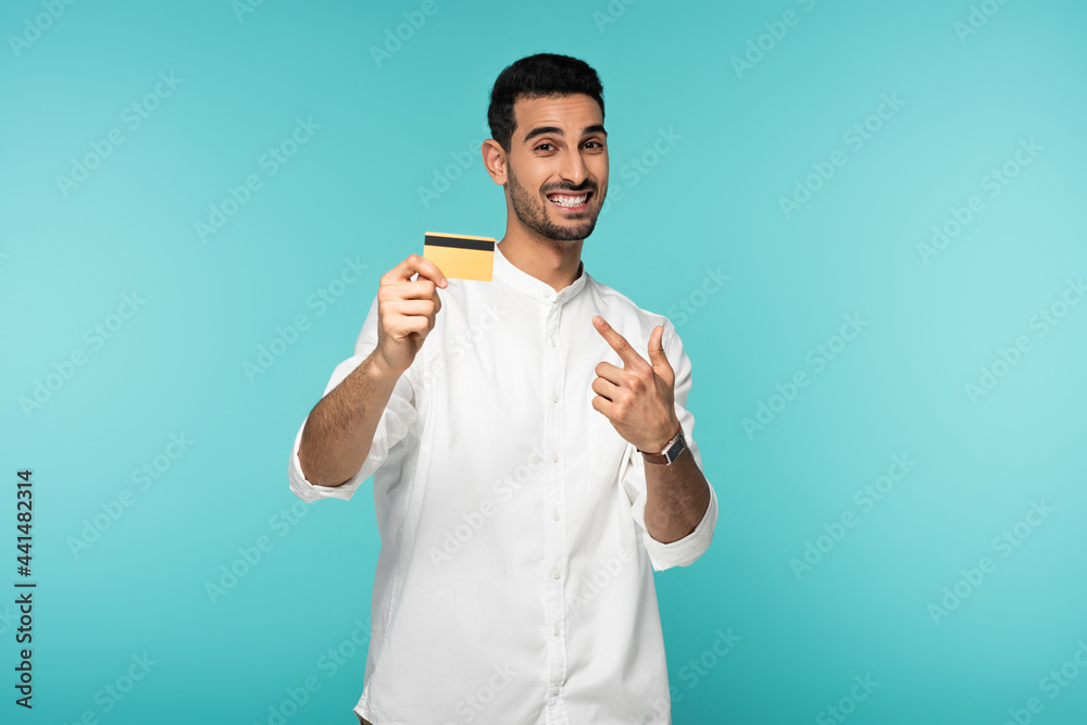 Cheerful arabian man pointing at credit card isolated on blue Cheerful arabian man pointing at credit card isolated on blue