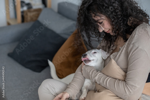 Young curly woman chatting surfing internet use smartphone on couch with dog Jack Russell Terrier