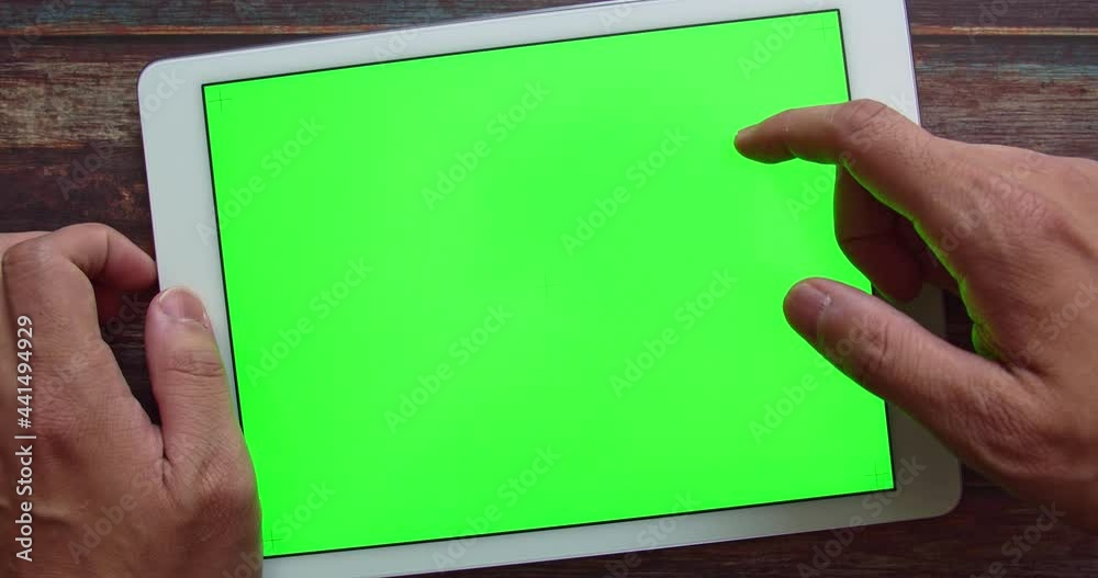 Man hands holding and scrolls and swipes Green Screen chroma key tablet on wooden shabby chic table. Mockup design at office or home in cozy mood. Dev tech, remote work and e-learning mock up concept.