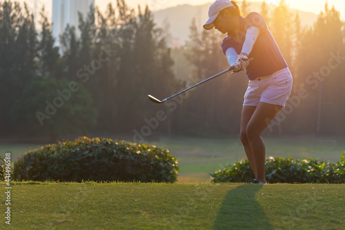 woman golf player in action of end downswing of wood driver, after hit the golf ball away from tee off to the fairway ahead, sunset scenery in background.