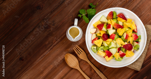 Mixed fruits salad including strawberry, kiwi, apple, and pineapple in white dish place on sackcloth on wooded table. Utensil and oil salad dressing cup beside with mint leaf decorating