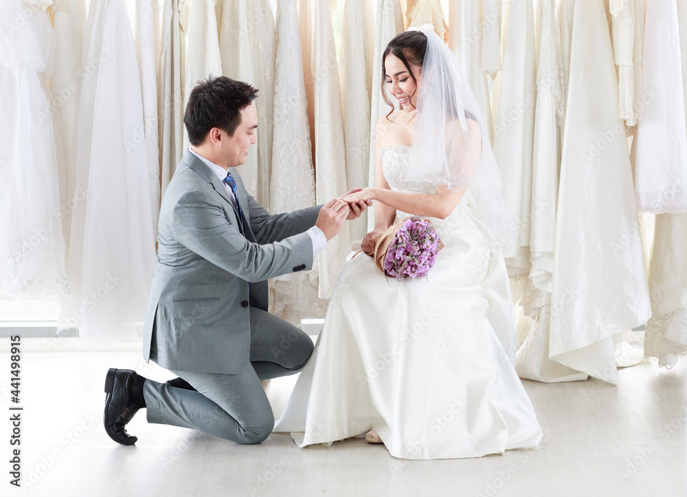 Fototapeta premium The groom in a gray suit kneels and wears a wedding ring to the bride in white lace in the room at studio. Asian couples having a blessed best day to share life. Concept happy lovely love.