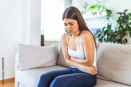 Woman in painful expression holding hands against belly suffering menstrual period pain, lying sad on home bed, having tummy cramp in female health concept