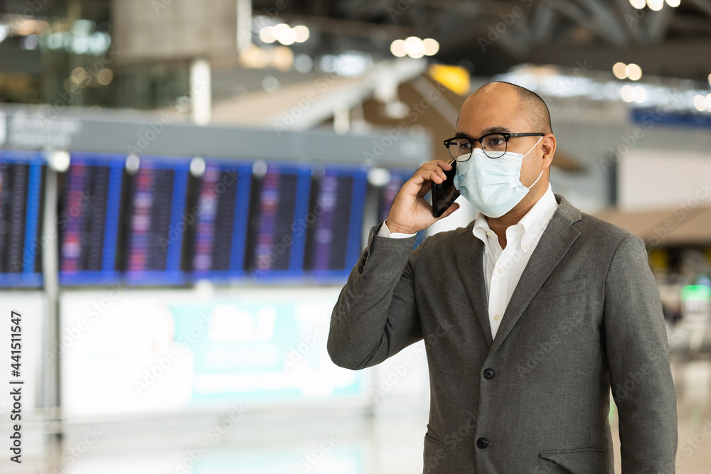 Young businessman talking on phone in the airport. Wearing mask. traveling during pandemic must be social distancing. He standing at information flight timetable waiting plane at airport terminal. Young businessman talking on phone in the airport. Wearing mask. traveling during pandemic must be social distancing. He standing at information flight timetable waiting plane at airport terminal.