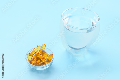 Bowl with fish oil capsules and glass of water on color background