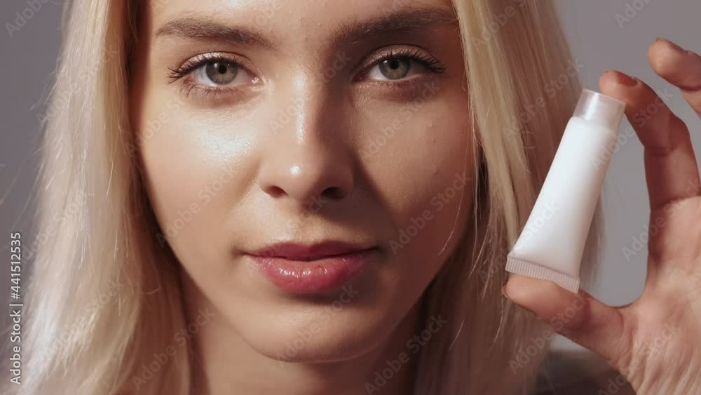 Skin moisturizing. Cosmetic product advertising. Facial care. Beauty wellness. Satisfied blonde woman with radiant face nude makeup showing white cream or lotion brand mockup bottle.