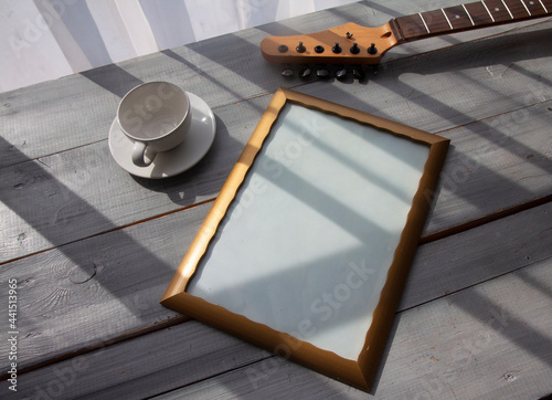 A frame for your lettering, a white cup for coffee or tea, a guitar neck on a gray wooden table, a sunny morning. The concept of congratulations, wishes, good morning.