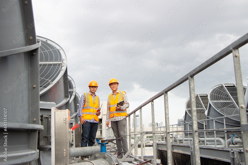 Asian engineer refrigeration plant  factory. male mechanic wearing helmet and Reflective Safety Vest uses laptop, tablet, equipment with sets of cooling towers conditioning systems background.