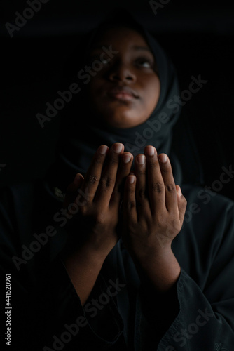 Dark skinned Muslim girl in black hijab, doing prayers on black background, selective focus