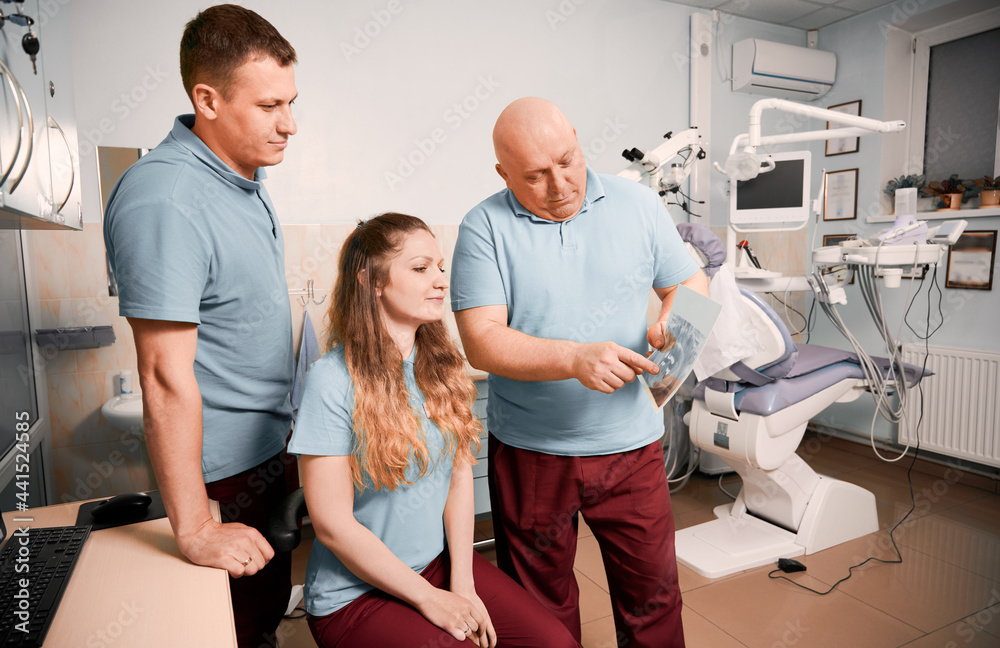 Obraz premium Male stomatologist pointing at dental scan while working with colleagues in dental office with modern equipment. Dentists team studying dental x-ray or orthopantomogram. Concept of dentistry.