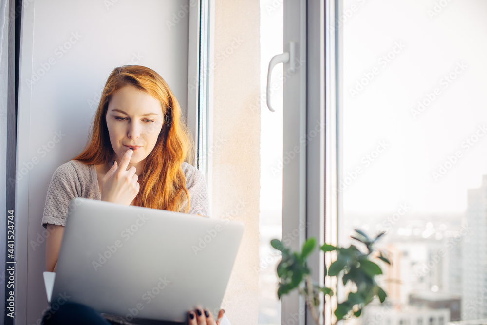 Young red-haired woman sitting at the window with laptop, using computer at home. Female freelancer in city apartment, selective focus, copy space. Young red-haired woman sitting at the window with laptop, using computer at home. Female freelancer in city apartment, selective focus, copy space.