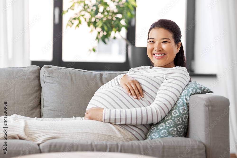 pregnancy, rest, people and expectation concept - happy smiling pregnant asian woman sitting on sofa at home pregnancy, rest, people and expectation concept - happy smiling pregnant asian woman sitting on sofa at home