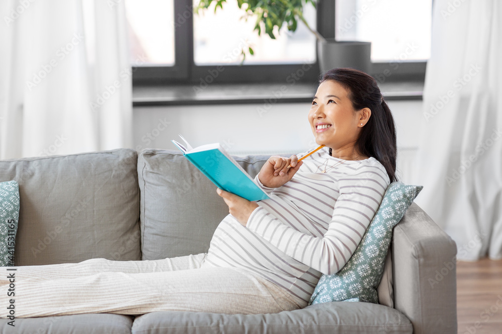 pregnancy, people and leisure concept - happy smiling pregnant asian woman sitting on sofa and writing to diary or notebook at home pregnancy, people and leisure concept - happy smiling pregnant asian woman sitting on sofa and writing to diary or notebook at home
