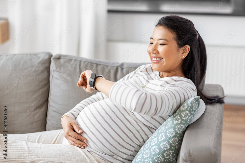 pregnancy, rest, people and expectation concept - happy smiling pregnant asian woman with smart watch sitting on sofa at home