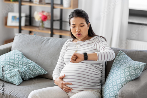 pregnancy, rest, people and expectation concept - pregnant asian woman with smart watch having labor contractions sitting on sofa at home and breathing photo