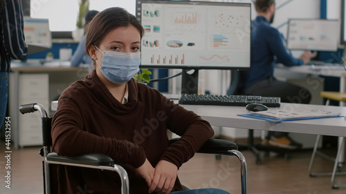 Woman employee with disabilities  invalid  handicapped paralized with protection mask against coronavirus looking at camera sitting immobilized in wheelchair in new normal business office room
