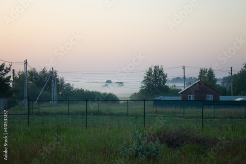 A village with a wooden house and a fence in the early foggy summer morning at dawn