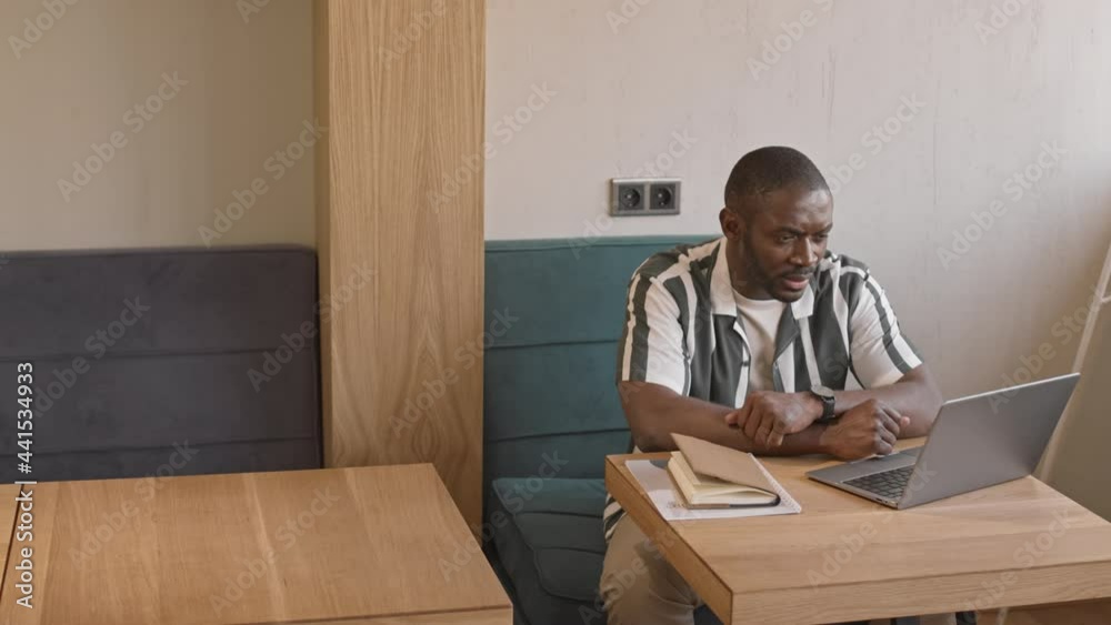 Medium shot of young smiling African-American man in casualwear video conferencing with friends or colleagues via laptop sitting at table in cafe Medium shot of young smiling African-American man in casualwear video conferencing with friends or colleagues via laptop sitting at table in cafe