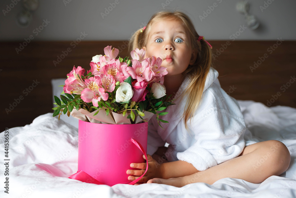 Little girl in a robe holds a box of flowers while sitting on the bed