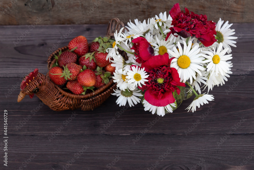 On a dark wooden background, there is a bouquet of daisies and poppies and ripe strawberries in a wicker dish.