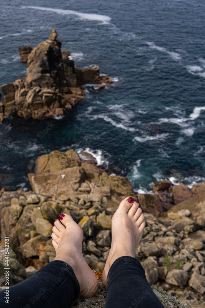 Photo of feet of a woman with red nails sitting on a rock in front of a beautiful landscape next to the sea. Amazing natural beach in UK. Lands End. Travelling in a sunny day