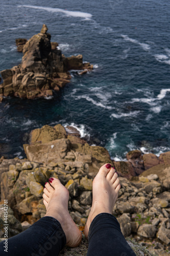 Obraz na płótnie Photo of feet of a woman with red nails sitting on a rock in front of a beautiful landscape next to the sea. Amazing natural beach in UK. Lands End. Travelling in a sunny day w salonie
