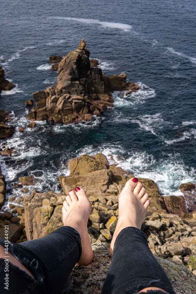 Photo of feet of a woman with red nails sitting on a rock in front of a beautiful landscape next to the sea. Amazing natural beach in UK. Lands End. Travelling in a sunny day