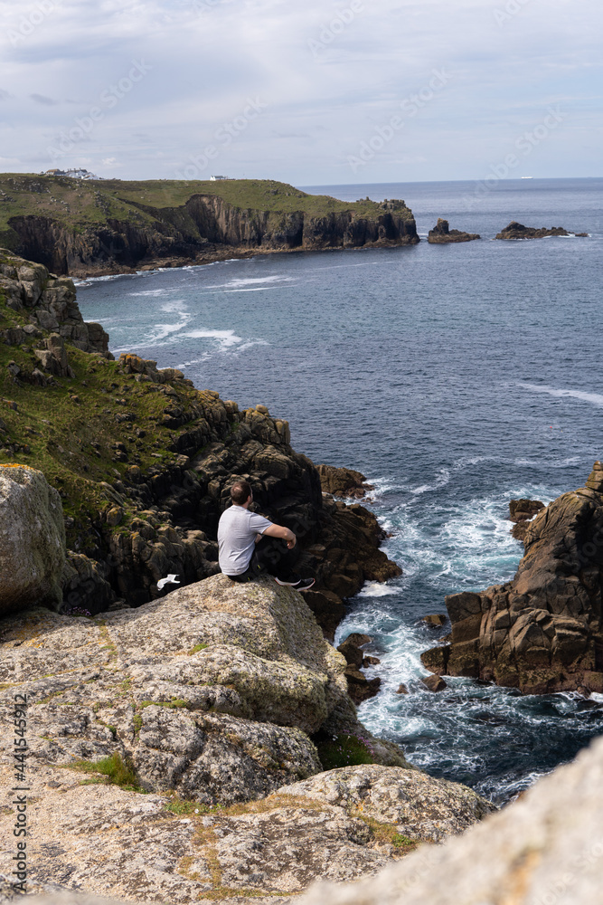 Photo of a young and attractive man sitting on a rock next to a cliff in the middle of nature, in Land's End, UK. Amazing landscape in a sunny day.